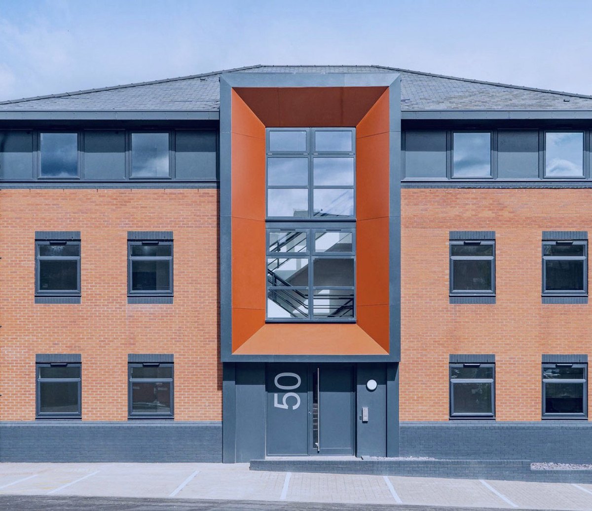 Photograph of the completed entrance to Sheldon Court, Birmingham: a tall orange clad bay projecting from the red brick elevation, with a full-height glazed panel above the dark grey entrance door and its painted number