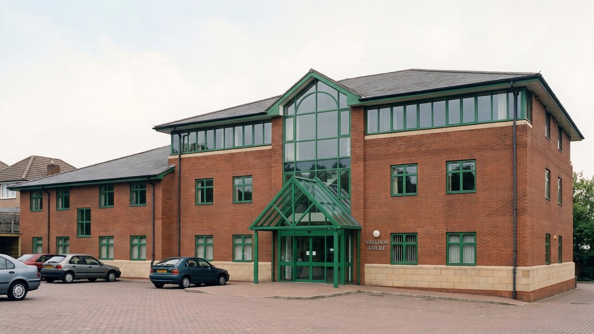 Visualisation of Sheldon Court in its office years: the same red brick blocks and grey hipped roofs, but with green-painted window frames, a tall green steel and glass gabled entrance porch, a buff stone plinth, cars parked on block paving and Sheldon Court signage beside the door
