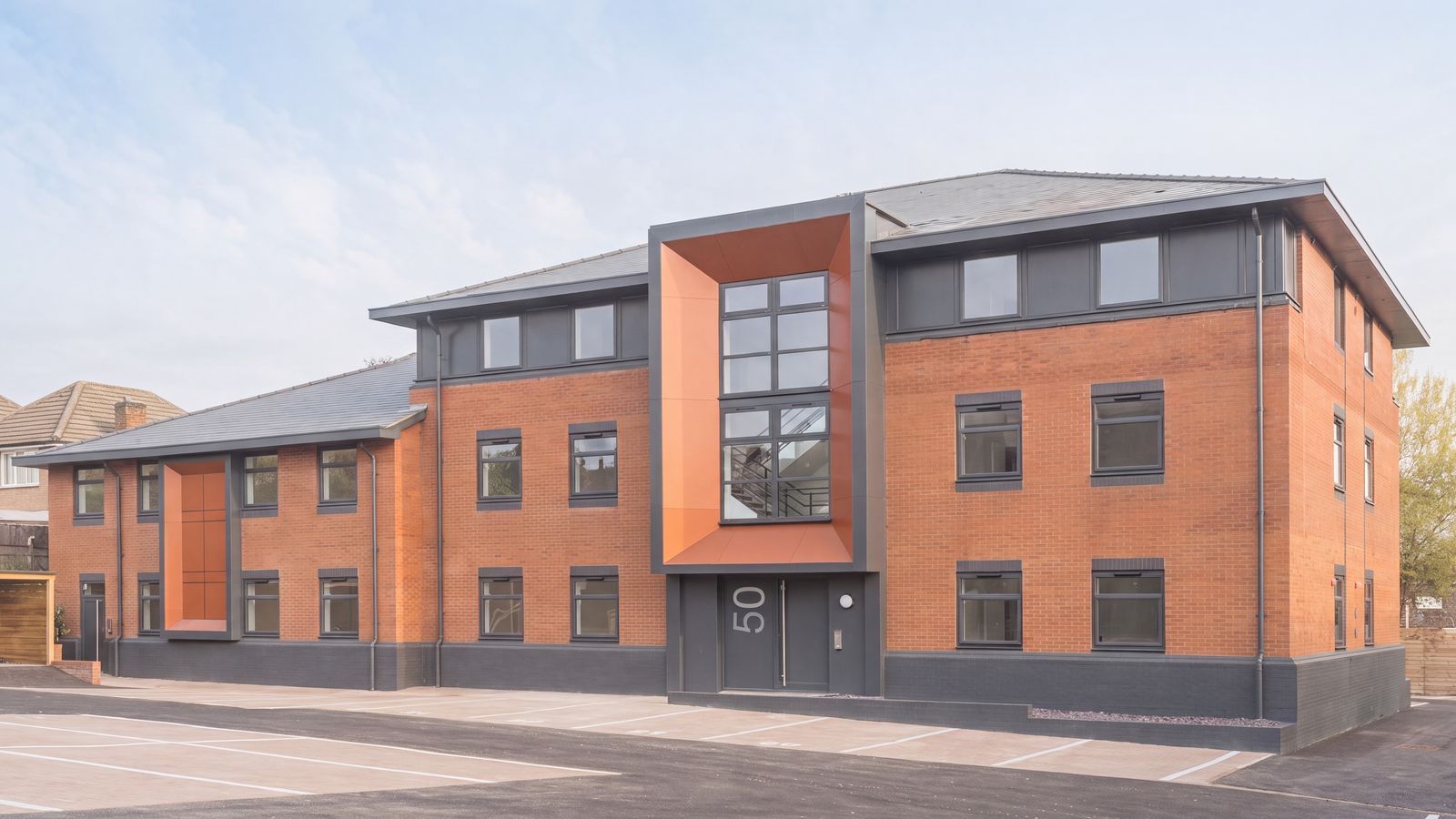 The completed Sheldon Court, Wagon Lane, Birmingham: two former office blocks converted to 64 apartments, in red brick with grey hipped roofs, anthracite window surrounds and a tall projecting orange entrance feature carrying the door number, with the resurfaced car park in front