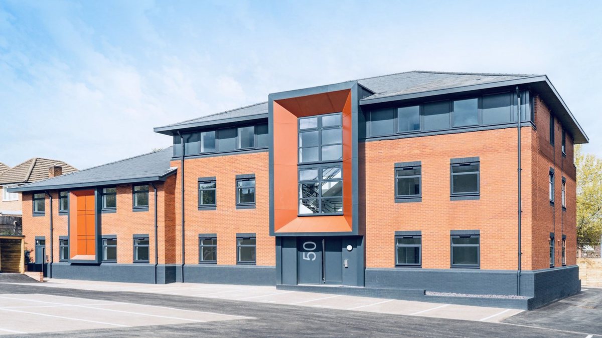 Photograph of the completed Sheldon Court from the same viewpoint: the identical brick blocks and roofs, now with anthracite grey window surrounds, a dark painted plinth, two projecting orange entrance features and a resurfaced car park