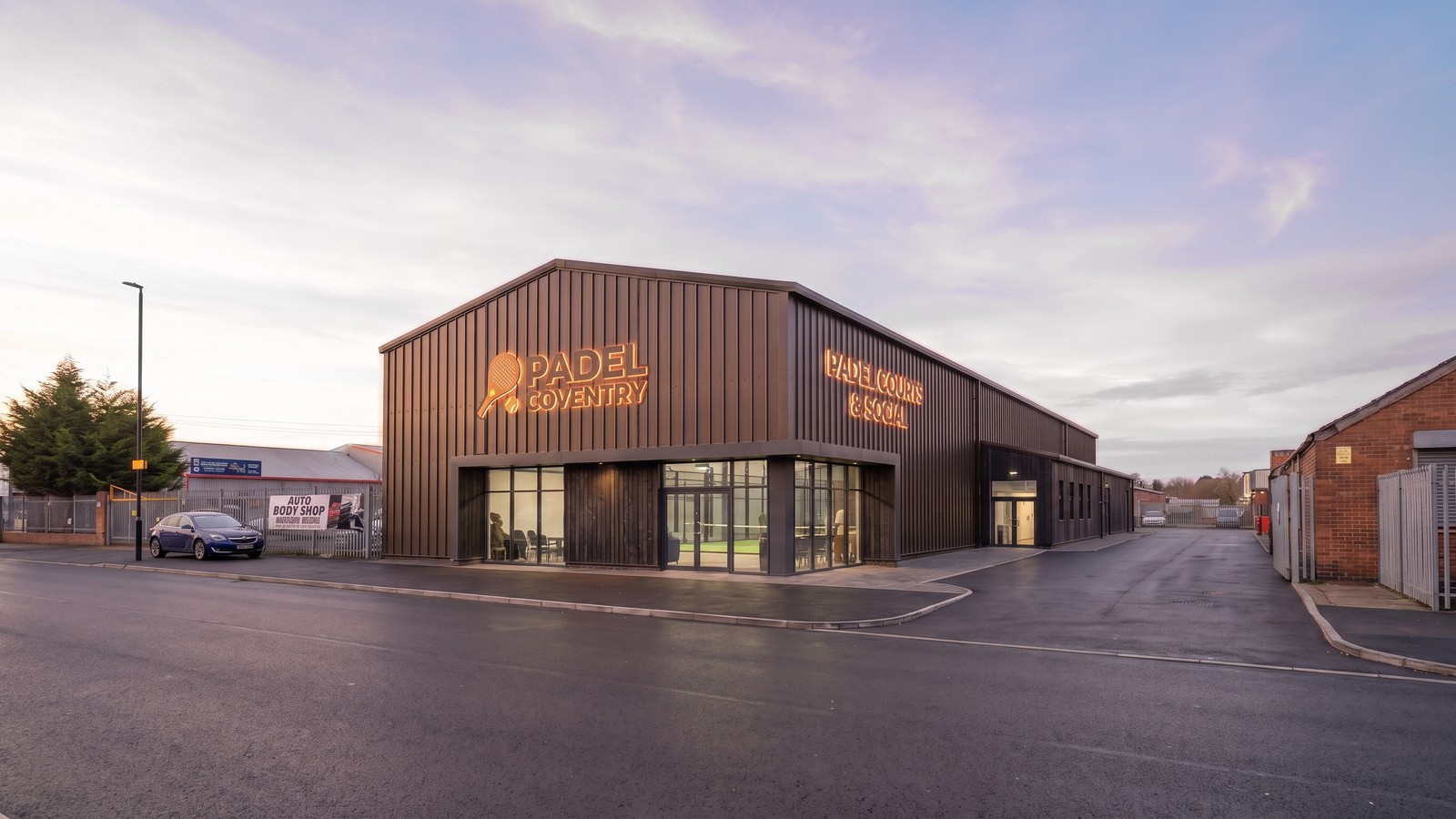 Visualisation of the proposed padel building at dusk: a long single-storey shed clad in dark vertical standing-seam metal, a shallow gable to the street with illuminated signage, and a fully glazed corner entrance showing the café and a court beyond, seen across wet tarmac inside an industrial estate