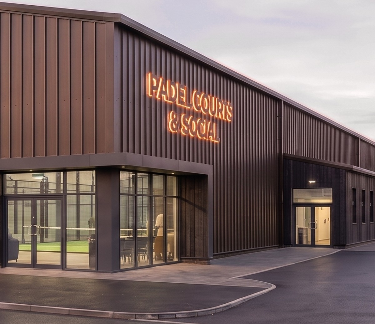The proposed building seen closer in: a glazed corner showing a padel court and the café behind the glass, dark standing-seam cladding above with illuminated fascia signage, and a second entrance door further along the flank