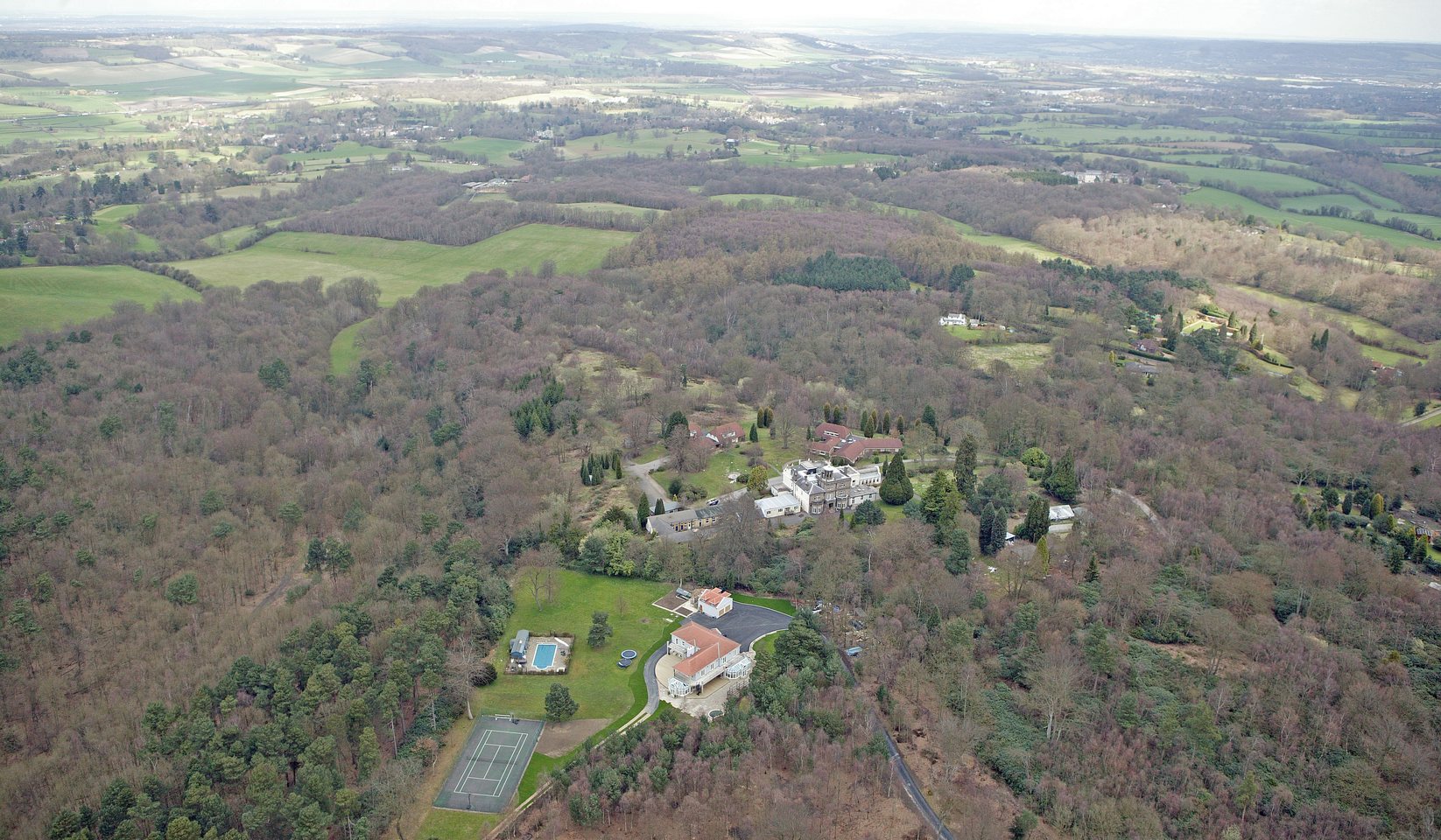 Aerial photograph of the Ide Hill site in 2006, before the development: the pre-existing buildings and grounds set in mature woodland on a ridge near Sevenoaks, with fields and open country stretching away beyond. None of the buildings shown are the eight houses