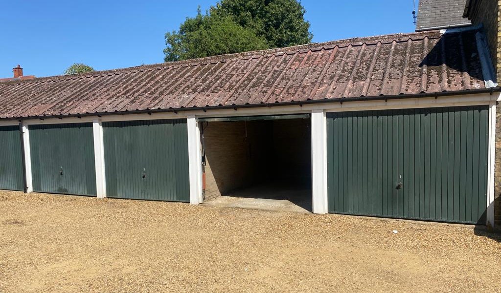 The run of seven garages at Burghley Mansions photographed in August 2022: green doors in white frames under a continuous pitched roof, one door open, gravel forecourt in front