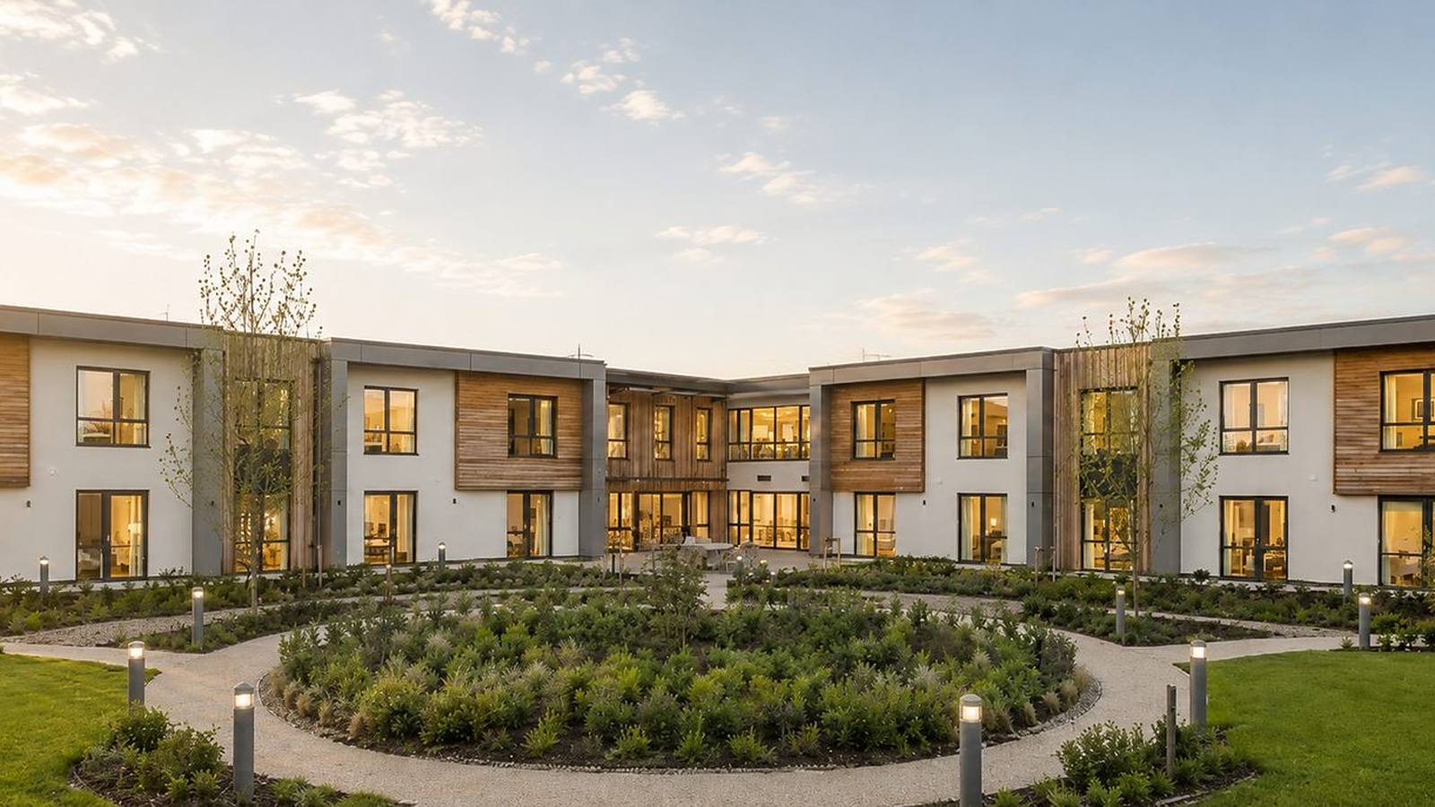 Visualisation of Brackley Care Home at golden hour: the two-storey home wrapping a symmetrical courtyard in white render with timber cladding and grey panels, its rooms lit warm from inside, a circular planted sensory garden in the foreground and lit bollards along the paths