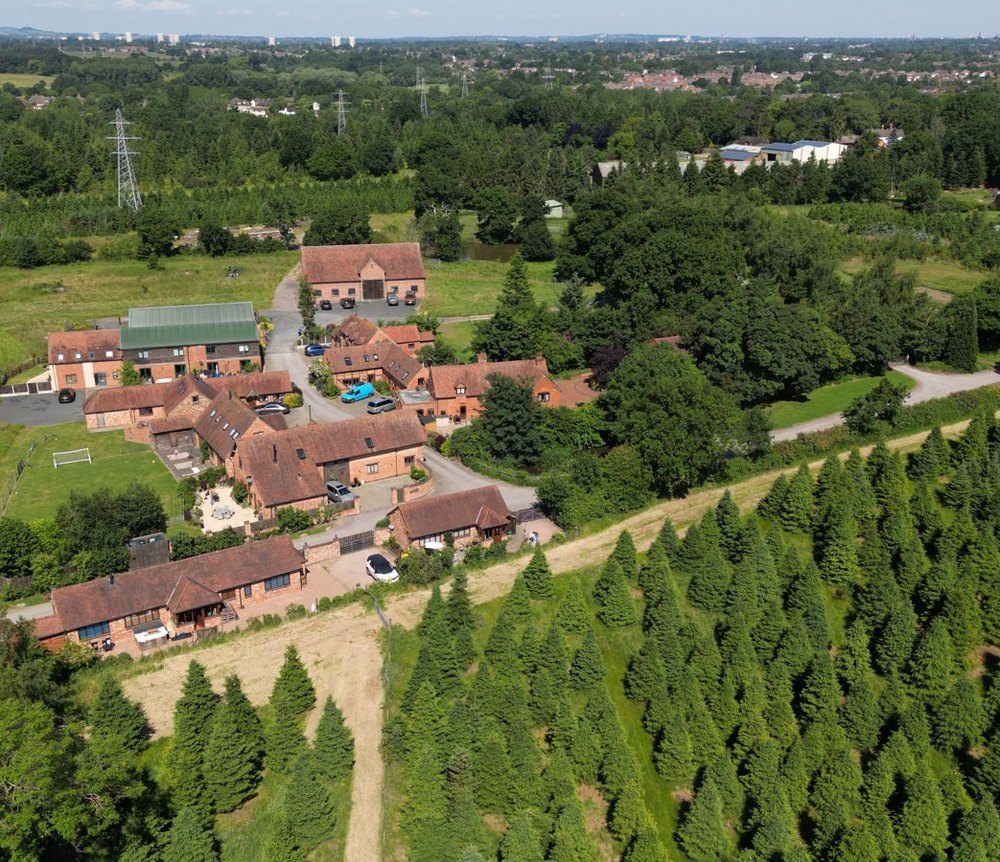 Aerial photograph of the Whitlocks Farmhouse cluster at Woods Farm: a group of brick farm buildings with clay tile roofs among mature trees, rows of Christmas trees running away to one side, and the Birmingham skyline on the far horizon