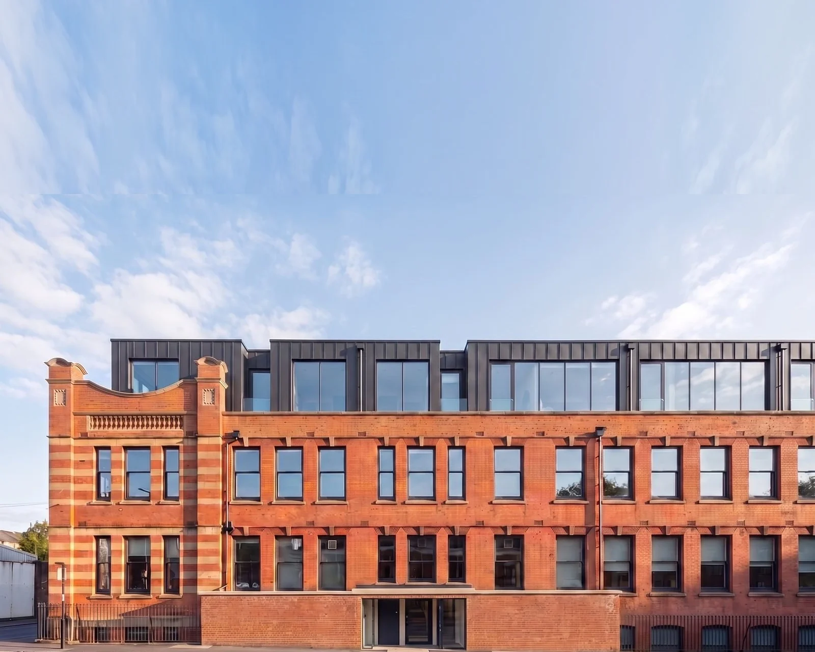 Enhanced photograph of the converted 1906 works at Foleshill Road, Coventry: a retained red brick and terracotta facade with an ornate gable, below a new dark zinc clad top storey. The sky above the roofline is extended.