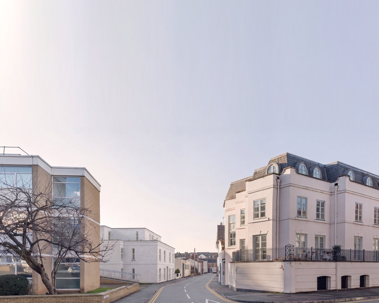 Photograph of the completed apartments at Cross Street, Leamington Spa: a three-storey building in white render behind a Regency terrace frontage, on a town-centre street. The sky above the roofline is extended.