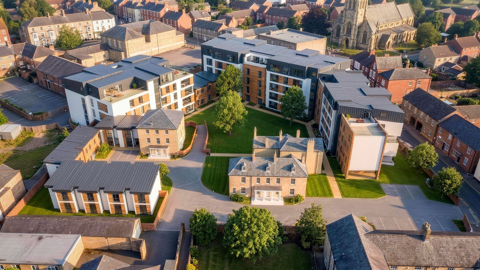 Aerial visualisation of the consented Caldecott Square scheme in Rugby: the retained Georgian listed building standing at the centre of a lawned courtyard, with new extra care apartment blocks in zinc, copper and timber ranged around the edges of the site, two-storey mews to one side, and the tower of St Andrews Church beyond