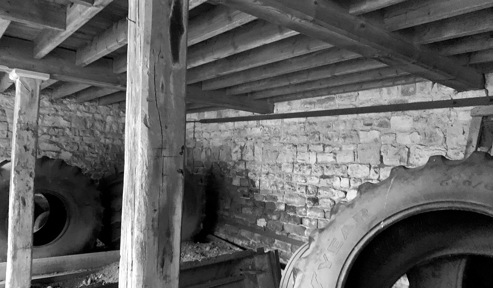 Black and white photograph inside one of the cart bays as found, looking up at the loft floor carried on close timber joists and a timber post, with the coursed stone and brick wall behind and tractor tyres stored under it