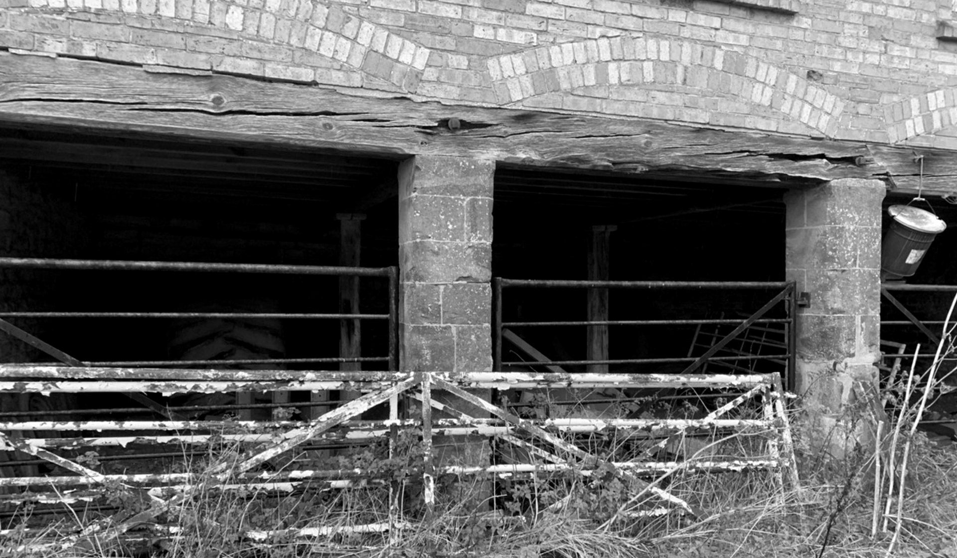 Black and white photograph of the cart house bay front as found: two of the four cart openings, divided by a dressed stone pier, headed by shallow brick arches under a heavy timber beam, with collapsed metal field gates standing across them