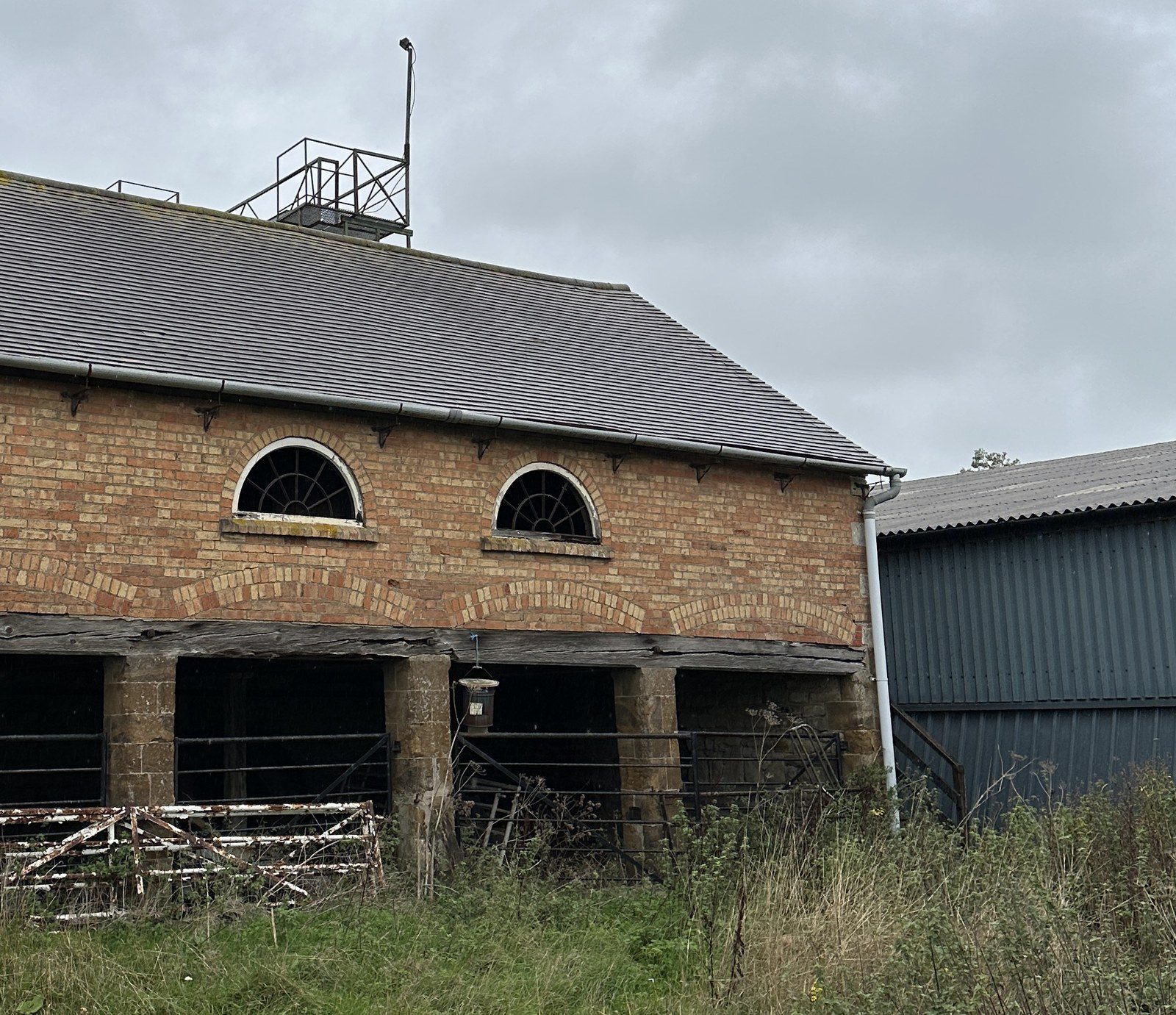 The cart house as found, in colour: four dark cart openings on stone piers under a timber beam, the brick wall above with two semicircular windows and its tiled roof, and the modern steel farm buildings pressing in on both sides