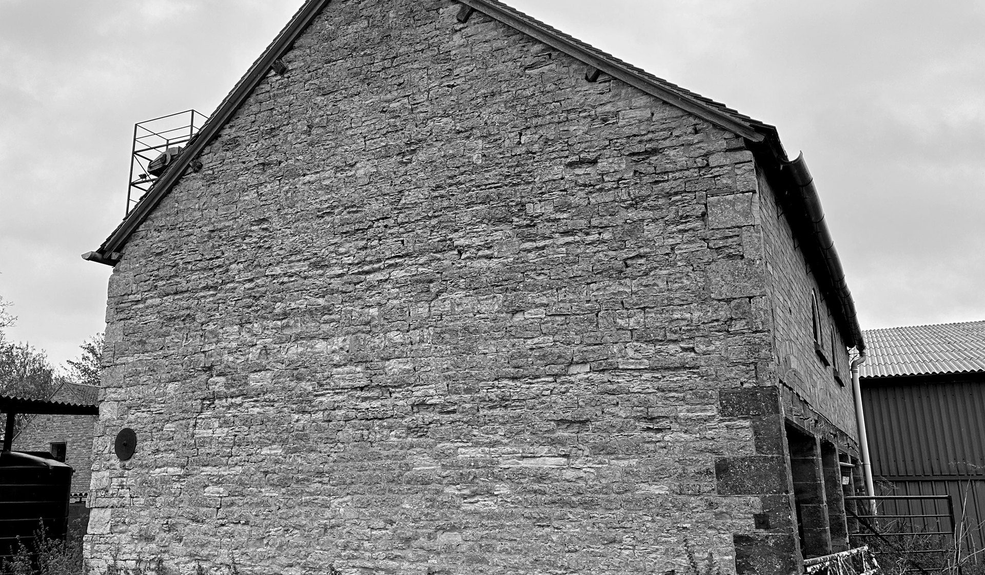 Black and white photograph of the cart house gable as found: a broad wall of coursed rubble limestone with dressed quoins and a small round opening, a weathervane at the apex, and a modern steel farm building alongside