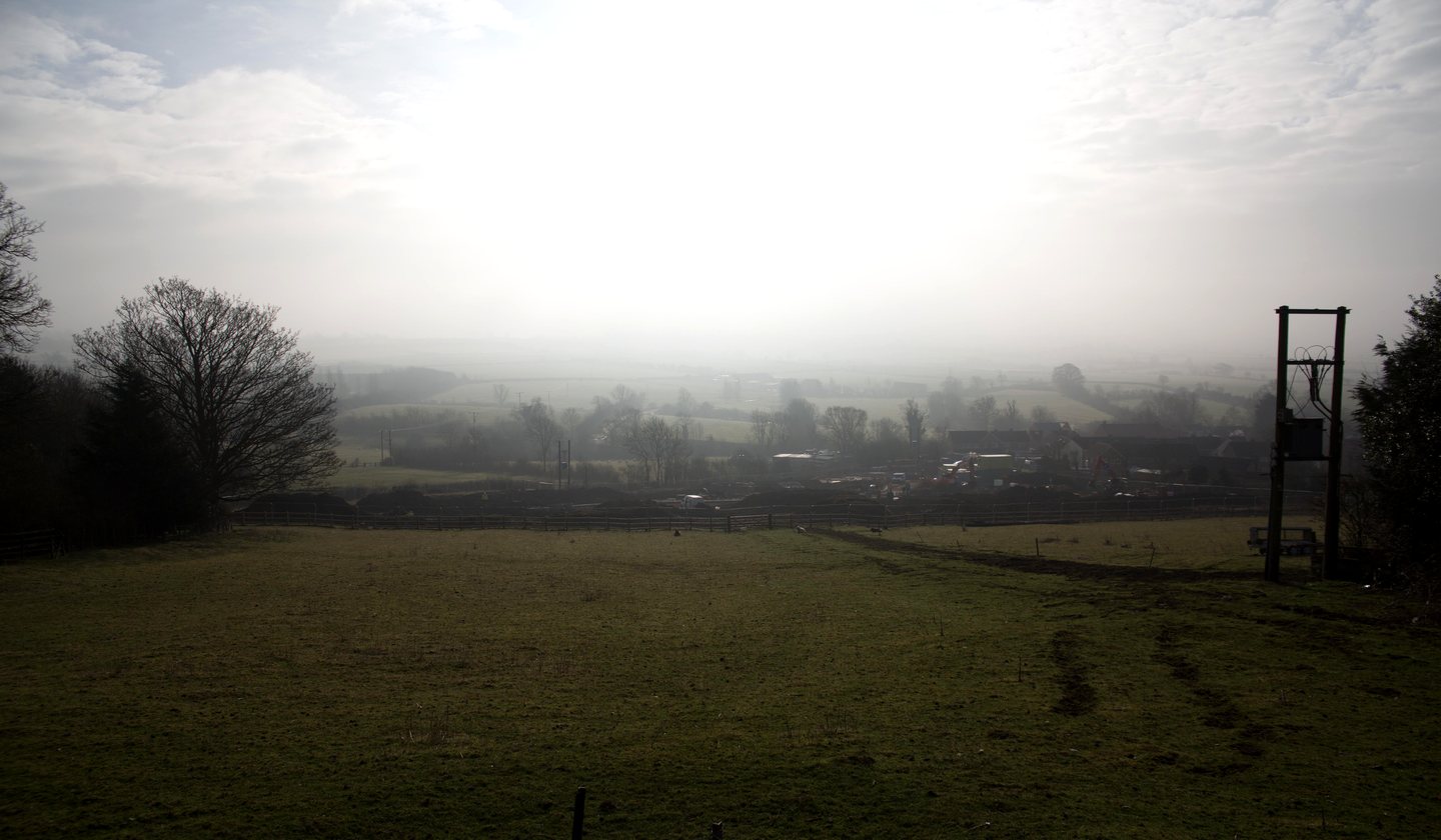 The view south from the site at The Woods, Napton-on-the-Hill in February 2018: pasture falling away from the hilltop to a wide misty vale of fields, hedgerow trees and the village edge below