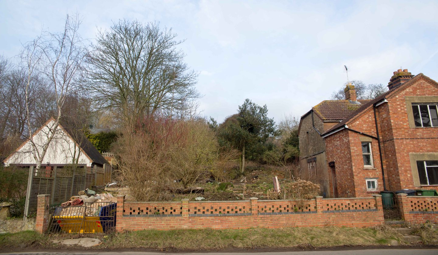 The plot at The Woods, Napton-on-the-Hill, photographed from Vicarage Road in February 2018: an overgrown garden strip rising behind a red lattice-brick boundary wall with an iron gate and a skip at its entrance, a white bungalow gable to the left and the flank of the brick cottage pair to the right