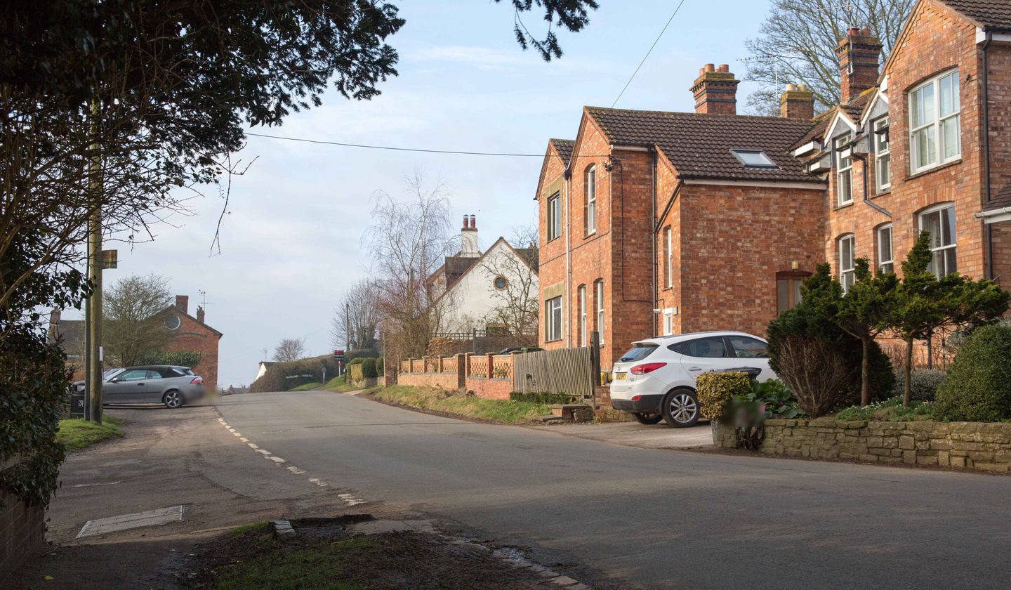 Vicarage Road, Napton-on-the-Hill, in February 2018, falling downhill beneath a dark yew, with the red brick cottage pair and a parked car on the right, the plot's lattice-brick wall just beyond, and a white cottage further down the lane