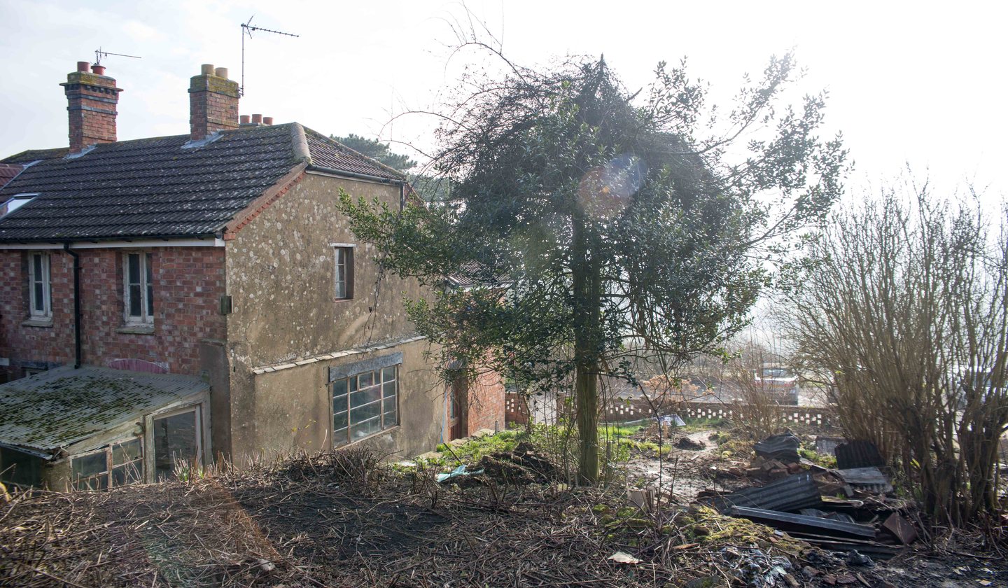 Looking back across the site at The Woods, Napton-on-the-Hill in February 2018: the rendered flank and rear of the red brick cottage pair with an old greenhouse lean-to, cleared scrub in the foreground, and the ground dropping to the lattice-brick wall and parked cars on Vicarage Road beyond