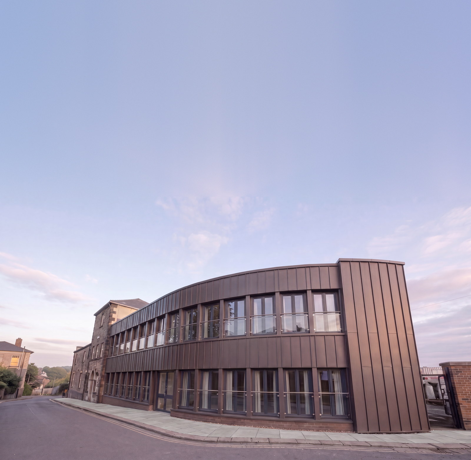 An indicative impression of 37 Station Road, Chesterfield as apartments: the retained stone range at the left and the curved frontage re-clad in bronze standing seam, seen from the station approach. The sky above the roofline is extended. Rickett Architects feasibility study.