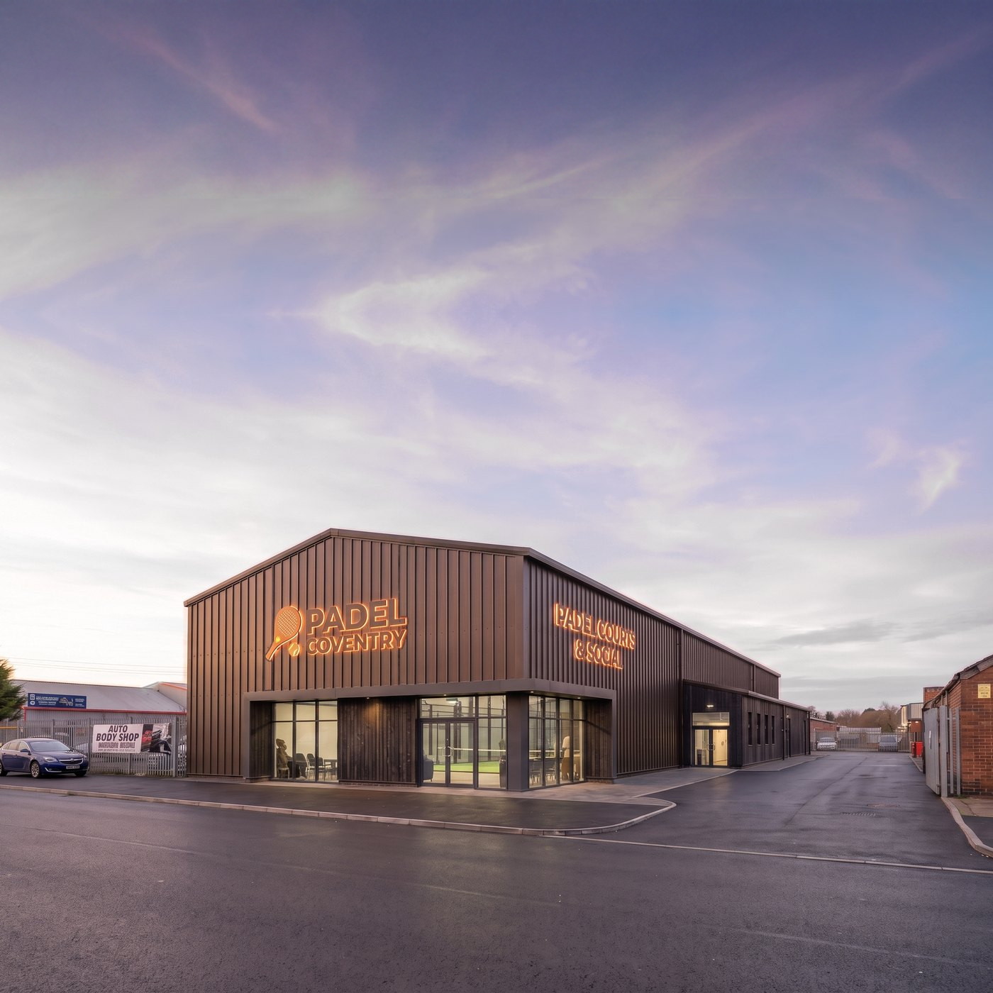 Visualisation of the proposed indoor padel building in Coventry at dusk: a dark timber-clad building with full-height glazing to the corner entrance, on an industrial estate.