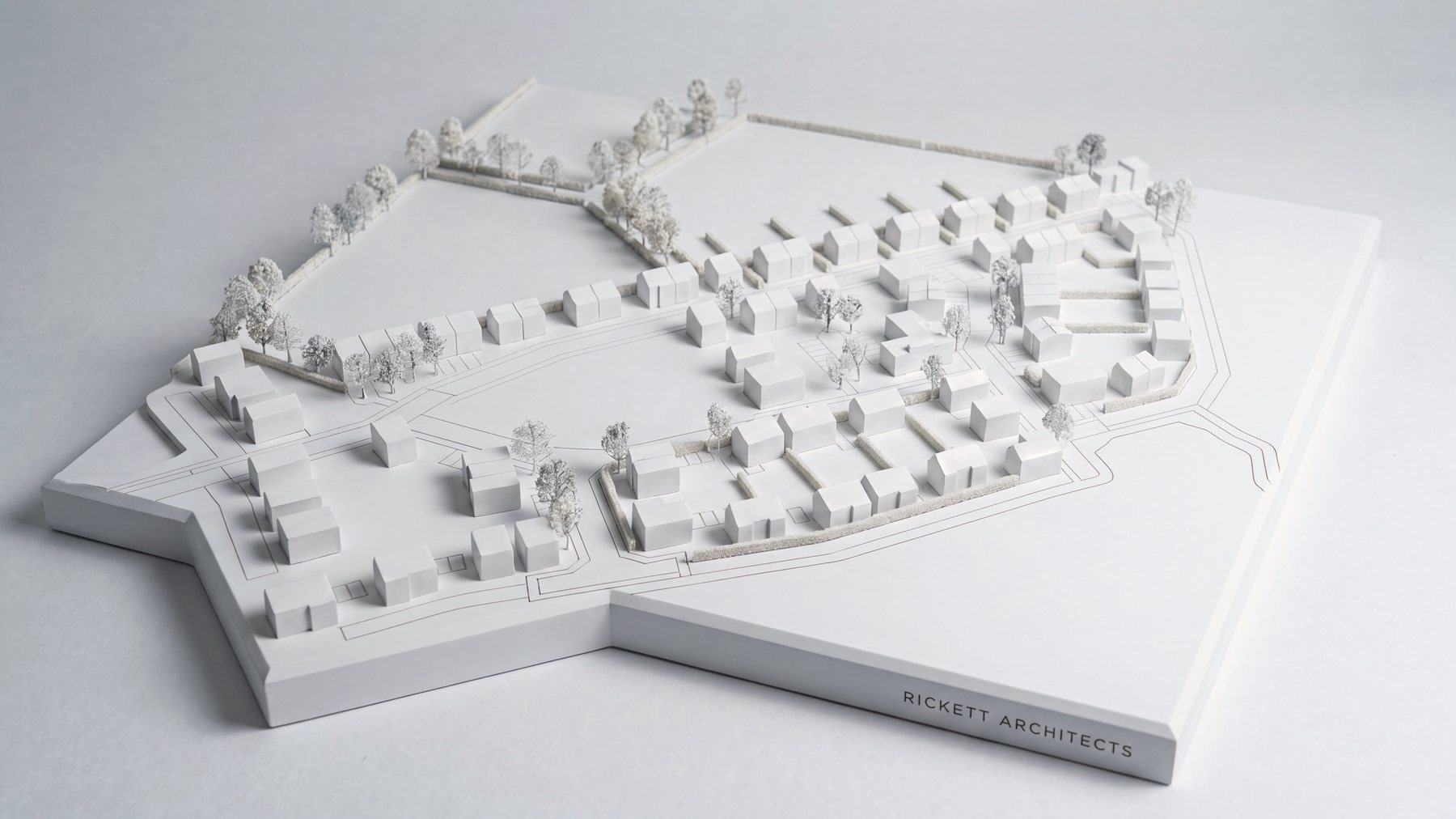 A white presentation model of the tested layout at Frog Lane, Balsall Common: ninety-five house forms in plain white laid out along curving streets across one half of a chamfered white plinth, with fine white model trees along the hedgerows, the open biodiversity field left empty on the other half, and the practice name engraved into the plinth edge