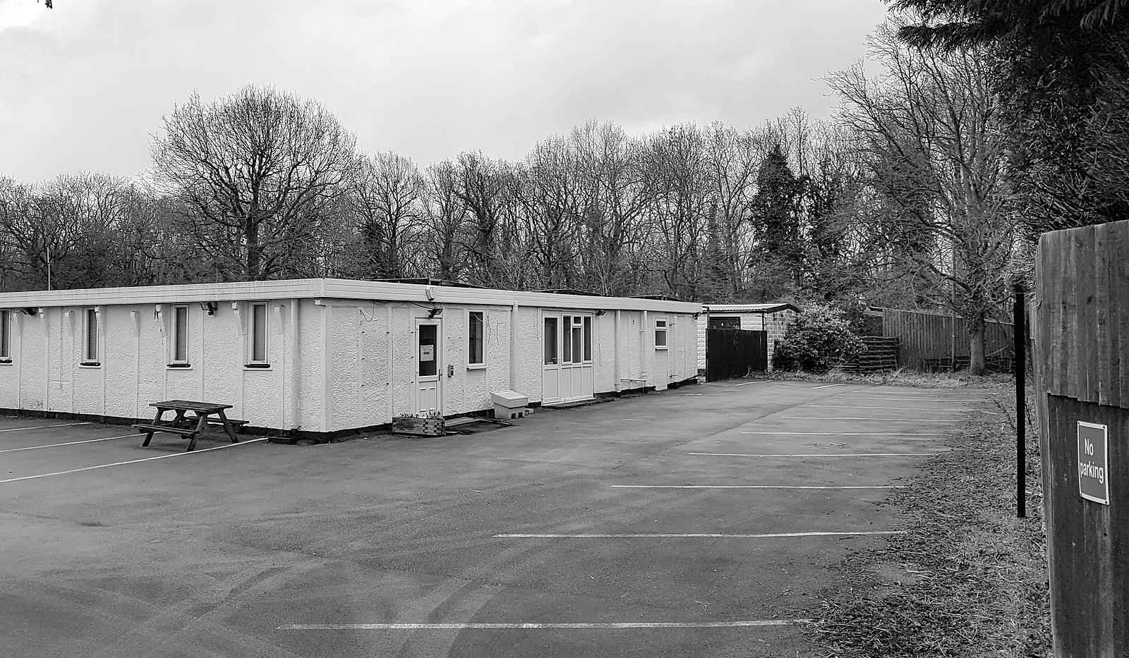 Black and white photograph of the site as found in March 2019: the long single-storey office range of the former air ambulance headquarters, painted white under a flat roof, standing behind a marked-out tarmac car park with a picnic bench, a close-boarded fence to the right and bare winter trees behind