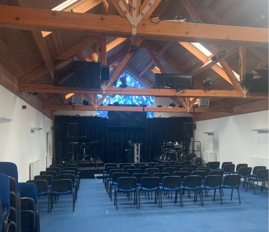 The church hall at 2 Priory Terrace, Royal Leamington Spa as found before the scheme: the hall looking towards its stage, with exposed timber king-post roof trusses, a stained-glass window above the stage, and rows of blue stacking chairs standing on the blue carpet floor beneath which the new basement was proposed