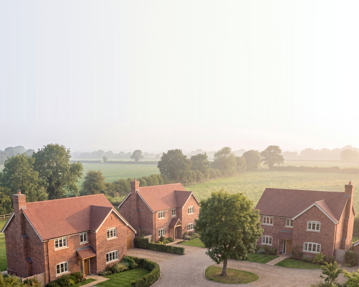 Visualisation of the consented scheme at Hob Lane, Burton Green: four two storey detached brick houses under clay tile roofs, arranged around a block paved shared drive with a retained tree at its centre, and open Warwickshire farmland beyond under an extended misty horizon.