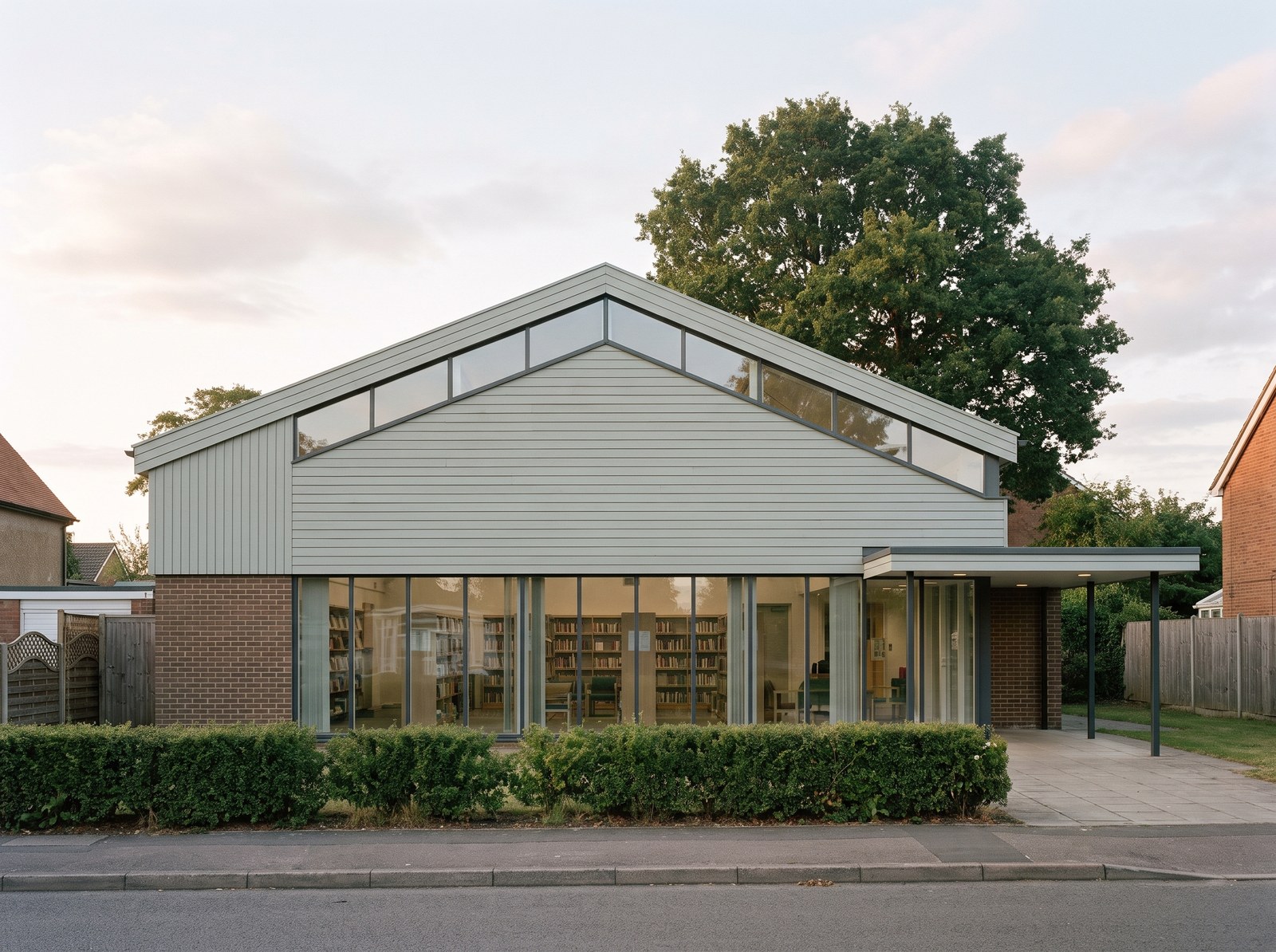 Illustrative view of Keresley library on Bennetts Road: a broad pitched gable clad in pale grey timber boarding with a band of clerestory glazing under the eaves, over a fully glazed ground-floor frontage with slender mullions showing the bookshelves inside, a flat-roofed entrance canopy to the right, a clipped hedge along the pavement and a large tree behind