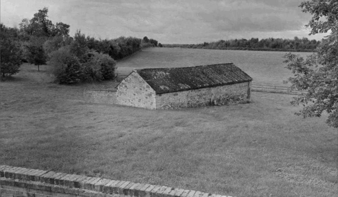 Black and white photograph of a long, low stone barn standing alone in a grass field, seen over a brick wall, with trees to the left and open fields running to a distant tree line