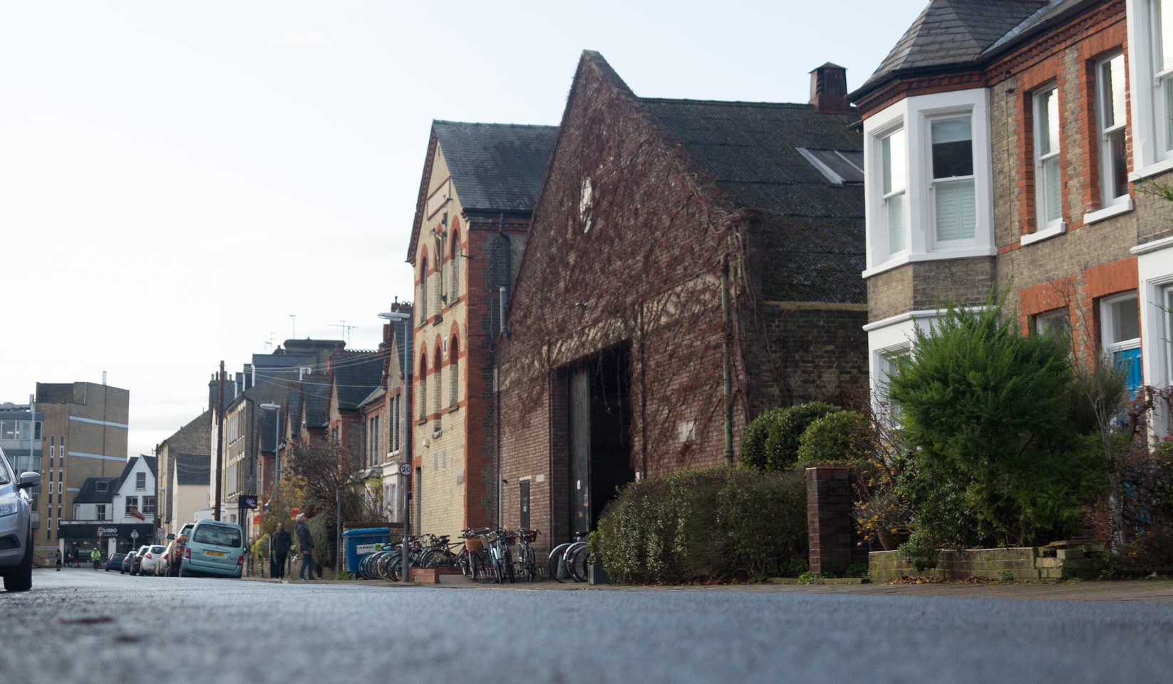 Photograph looking along the street in December 2019: a tall brick dance school building with arched windows, an ivy-covered brick warehouse with a pitched gable beside it, and bay-fronted brick houses in the foreground