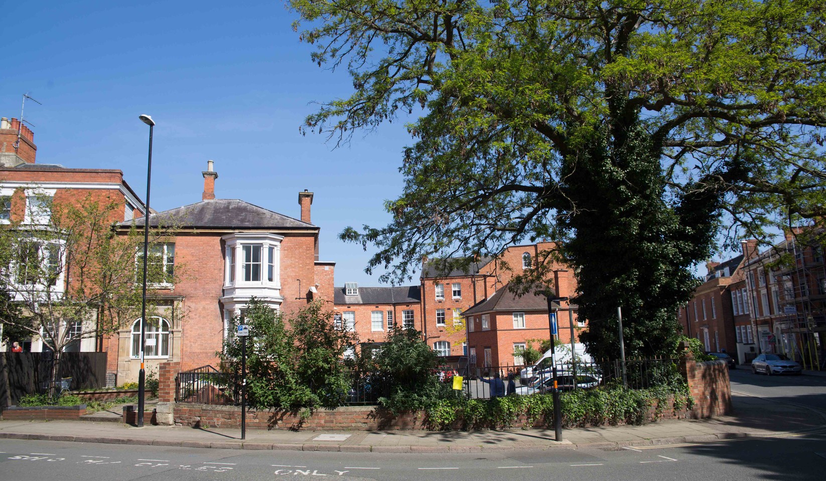 Photograph of a street corner in Northampton in bright sunshine: a red brick Victorian house with a white bay window behind a low wall and railings, a large tree in full leaf, a car park with a white van behind the railings, and Victorian terraces along the street beyond