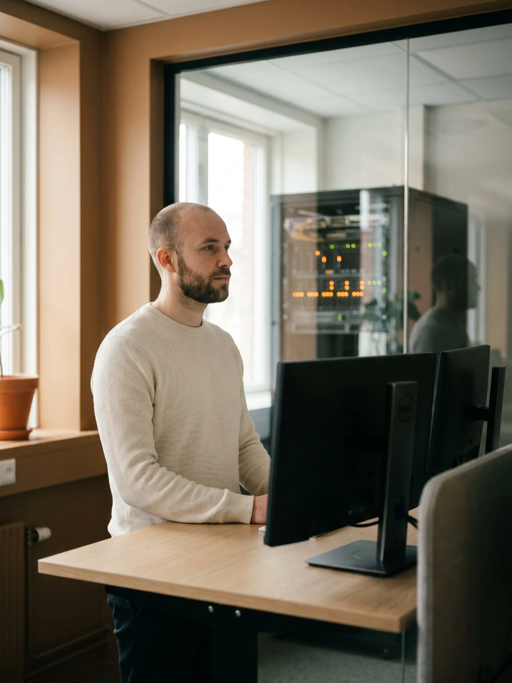 Developer at a standing desk beside a glass-walled rack of network equipment