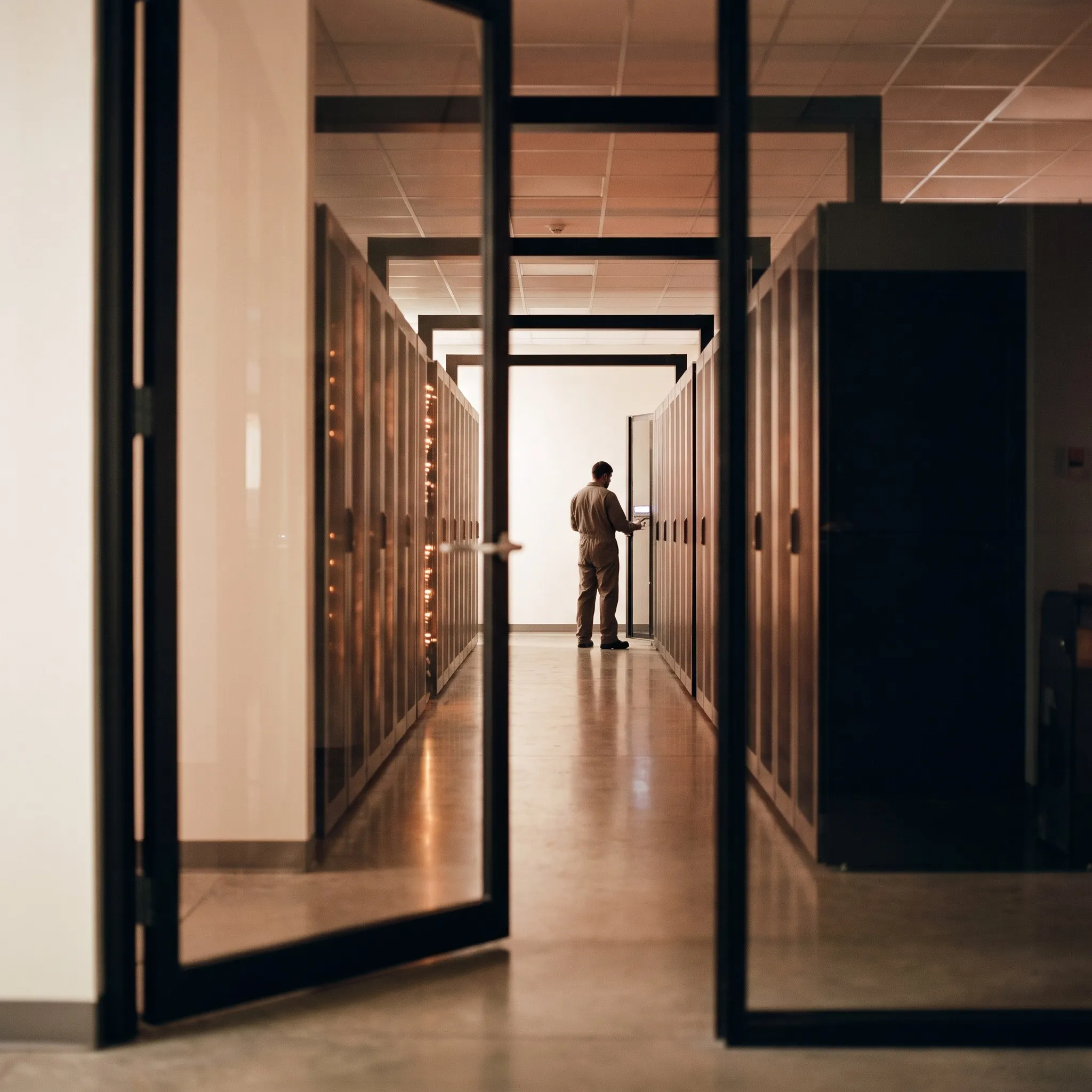Server room aisle seen through a glass door, a technician at the far end