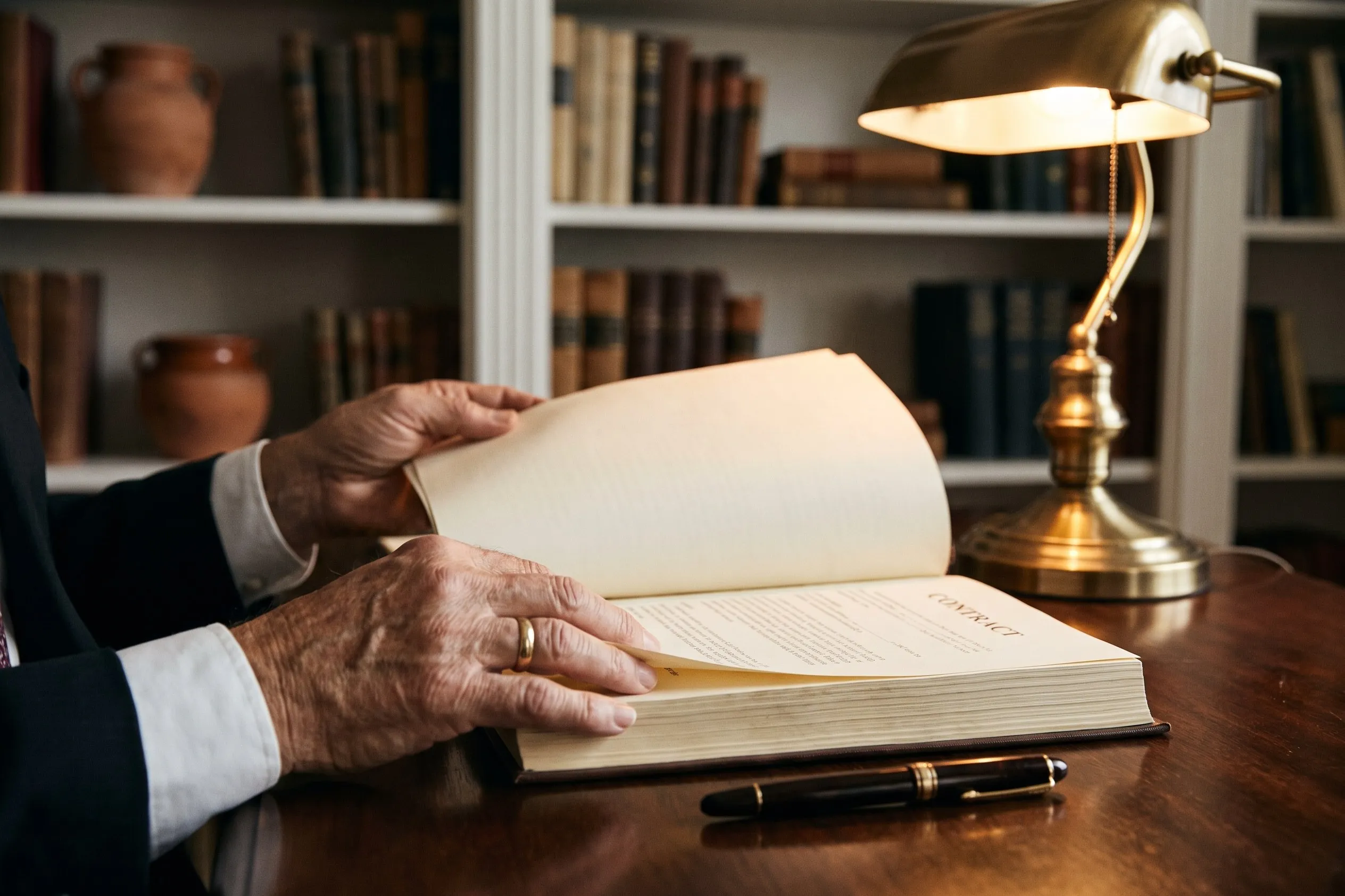 Hands turning the pages of a contract beside a fountain pen on a dark wood desk
