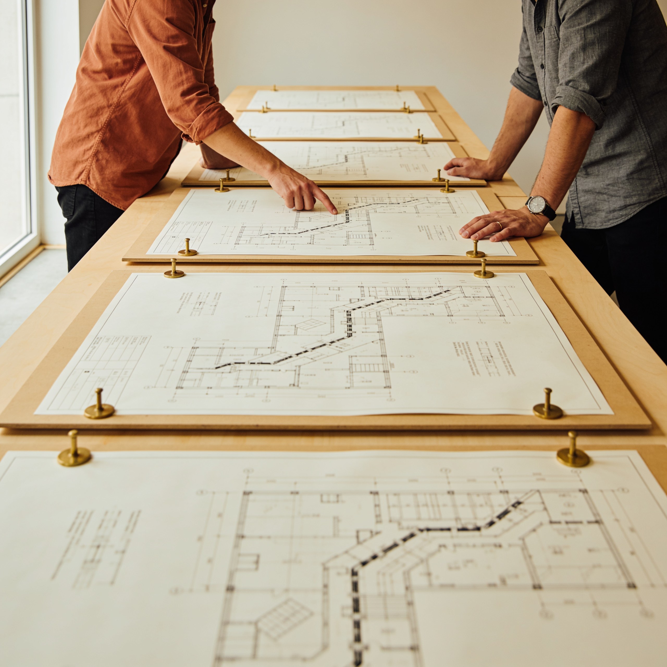 Two colleagues tracing a route across large printed engineering schematics held flat by brass weights