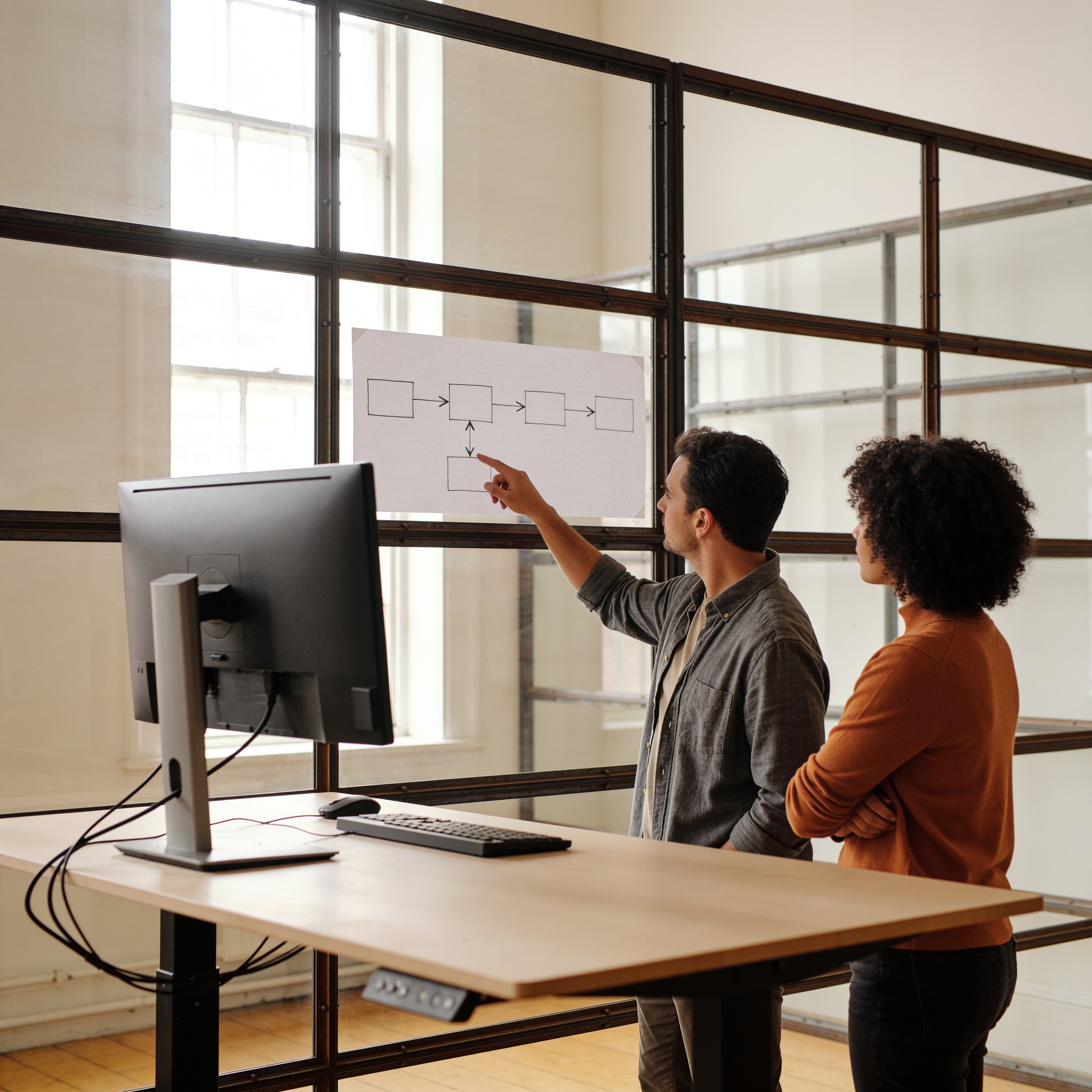 Two colleagues at a standing desk working through a printed process flow taped to a glass partition