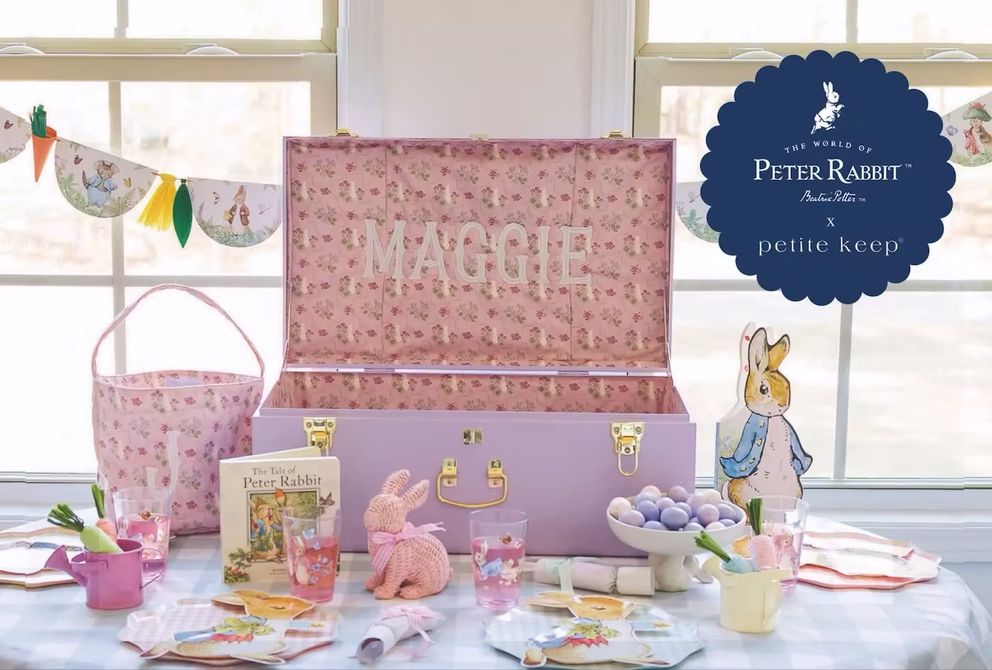 Petite Keep Postcard by PostPilot featuring collaboration trunk of Peter Rabbit and Petite Keep. The trunk is a light purple, opened, and says "Maggie" in subtle light pink lettering inside. In front of the trunk features an array of Peter Rabbit products and Easter items (e.g., dyed eggs).