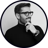 Man with glasses and beard wearing a black turtleneck, smiling and resting his chin on his hand.