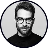 Portrait of a man with short dark hair, beard, glasses, and a black turtleneck sweater against a gray background.