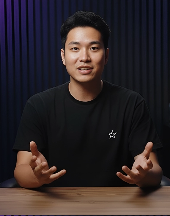 Young man with black hair and black t-shirt speaking and gesturing with hands at a wooden table against a dark curtain background.