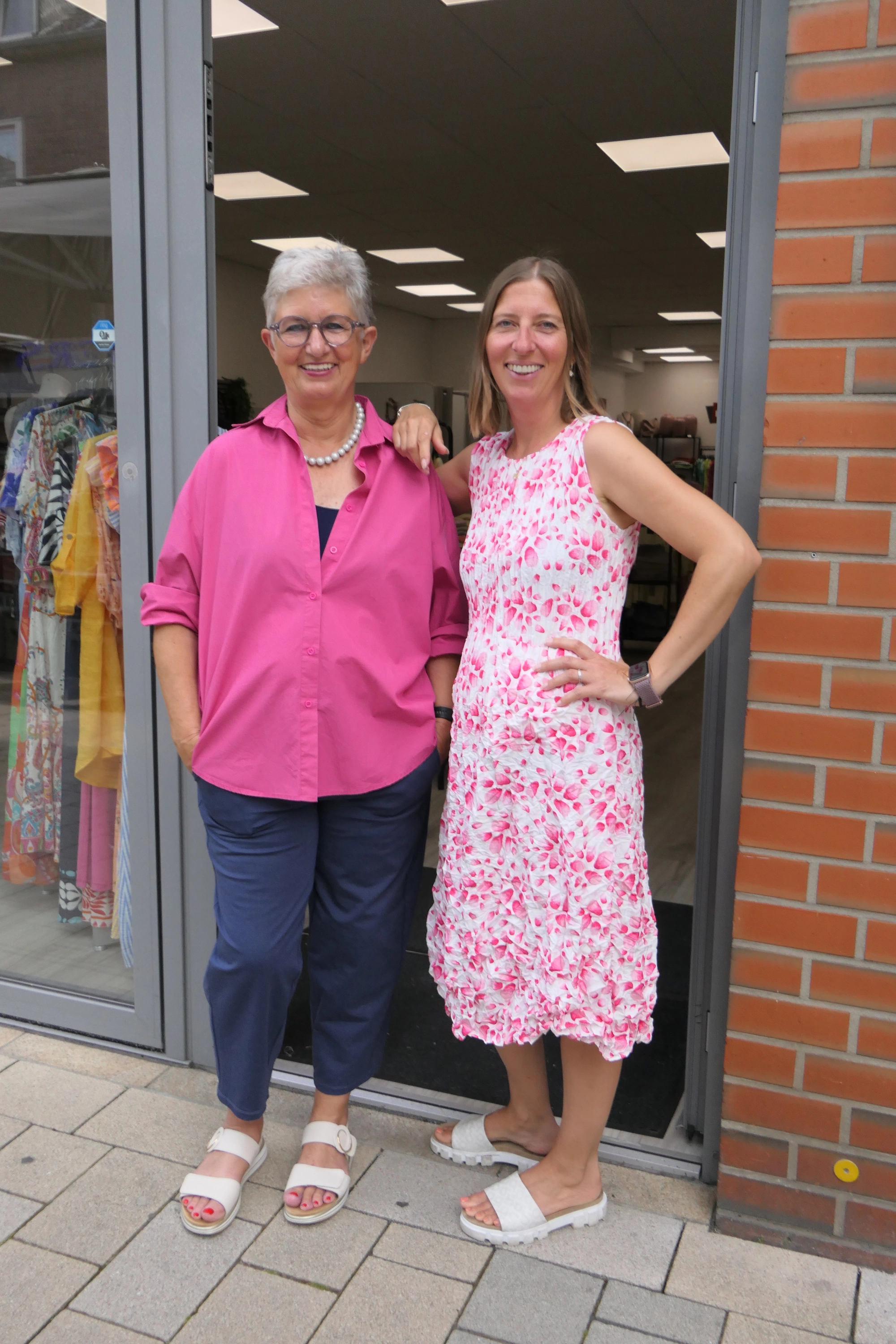 Zwei lächelnde Frauen - Marion Day und ihre Tochter - stehen nebeneinander vor dem Geschäftseingang von my day dress, eine trägt eine pinke Bluse und blaue Hosen, die andere ein weißes Kleid mit pinkem Blumenmuster.