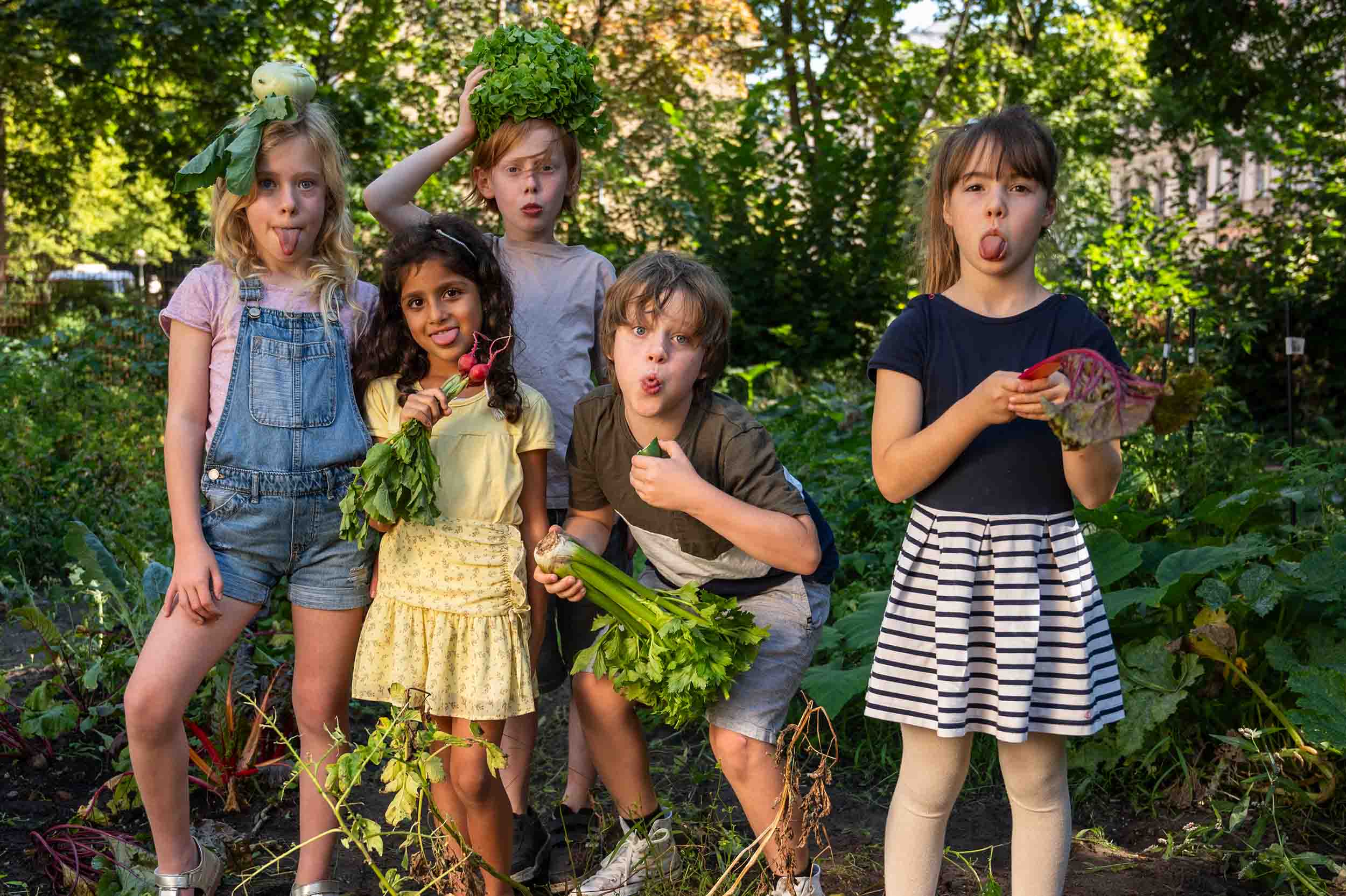 Fünf Kinder machen Grimassen im Garten, einige mit frischem Gemüse in der Hand oder auf dem Kopf.