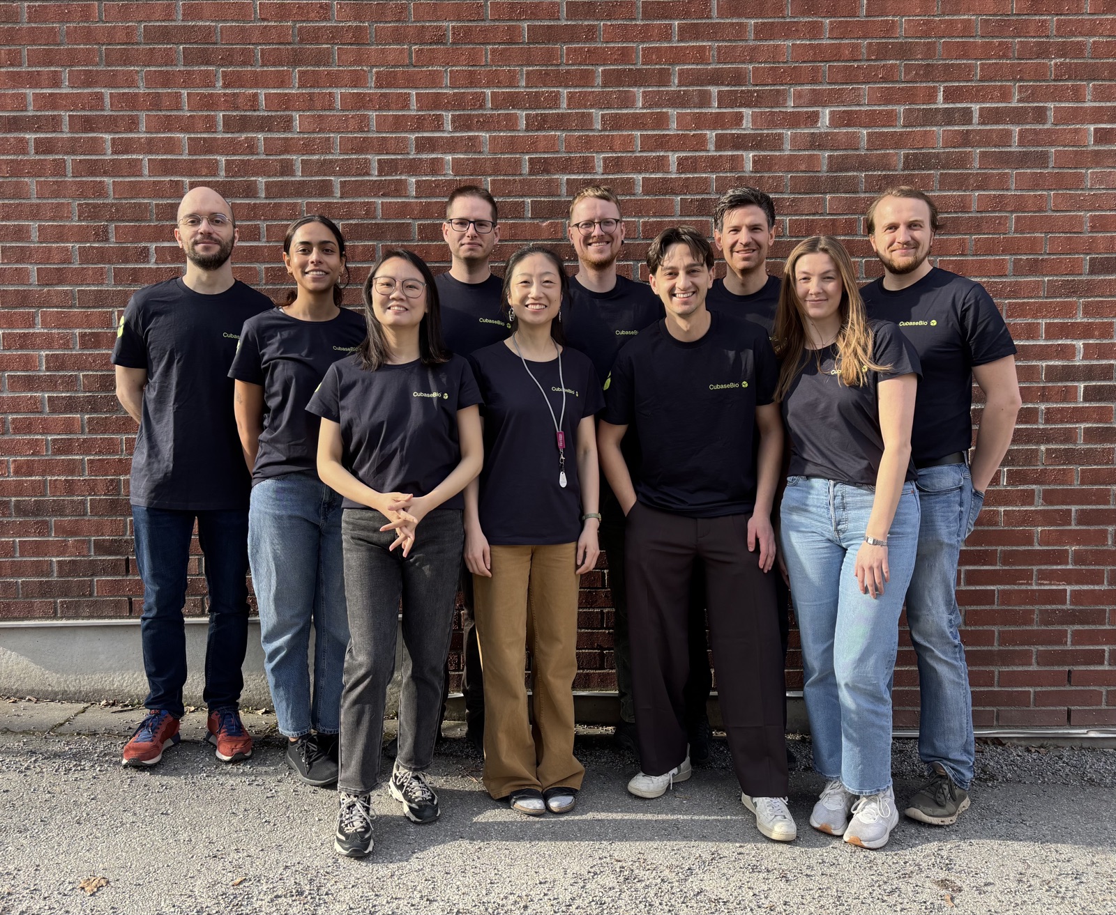 The whole Cubase Bio team standing in two rows in front of a brick wall, all wearing black t-shirts with a small green CubaseBio logo on the left chest, smiling towards the camera.