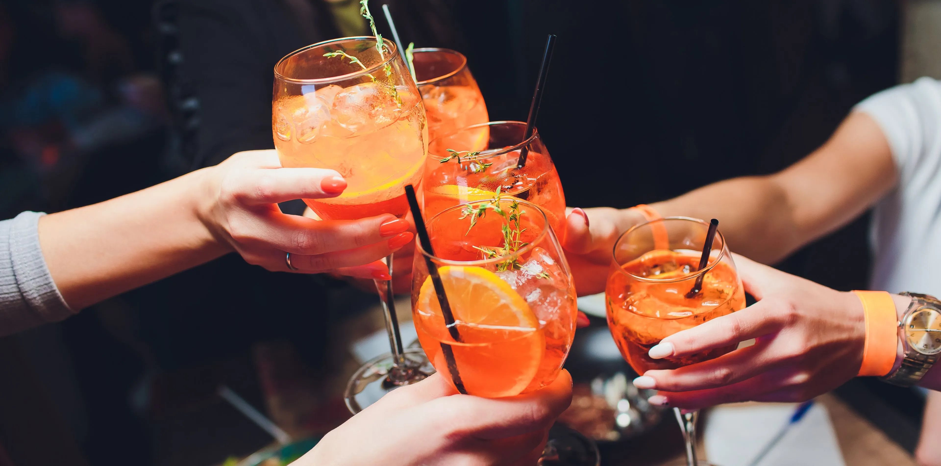 Des mains portant des verres de cocktail orange garnis de tranches d'orange et de thym.