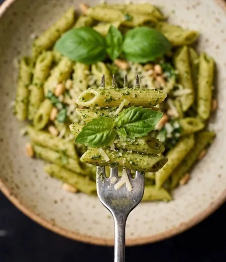 Fourchette tenant des pâtes penne au pesto garnies de feuilles de basilic et de pignons.