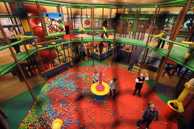 Kinder spielen in einem bunten Indoor-Spielplatz mit Bällen, Rutschen und Kletternetzen.