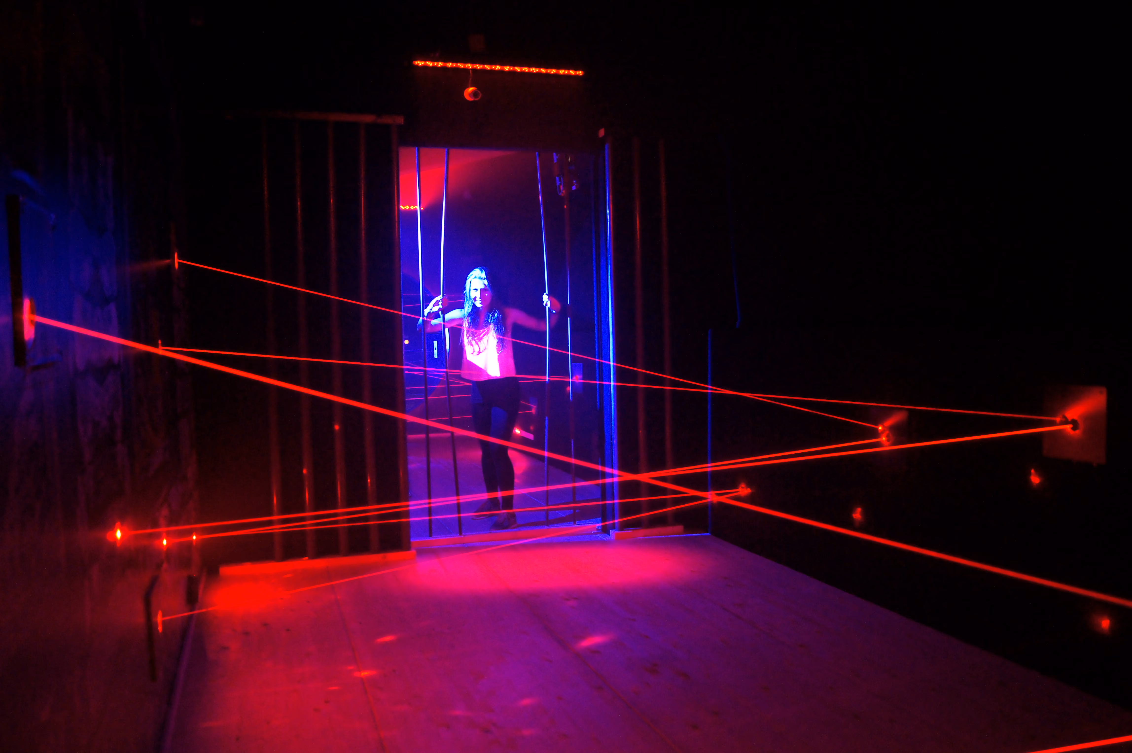 Frau bewegt sich vorsichtig durch einen Laserparcours mit roten Lichtstrahlen in dunklem Raum.