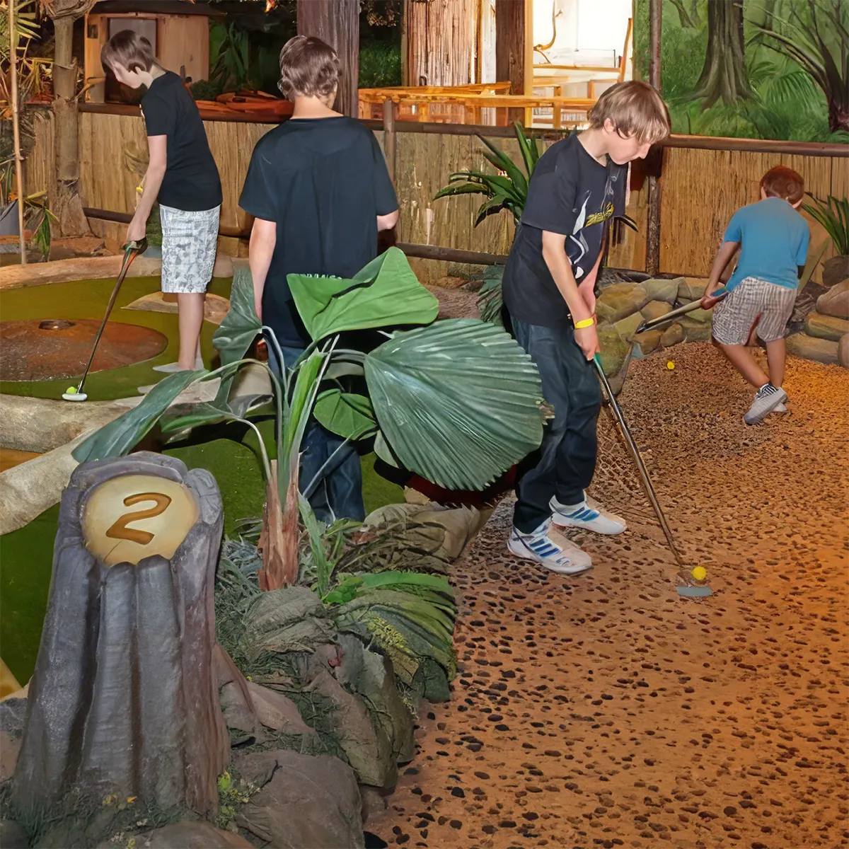 Vier Jungen spielen Minigolf in einer tropischen Indoor-Anlage an Bahn Nummer 2.
