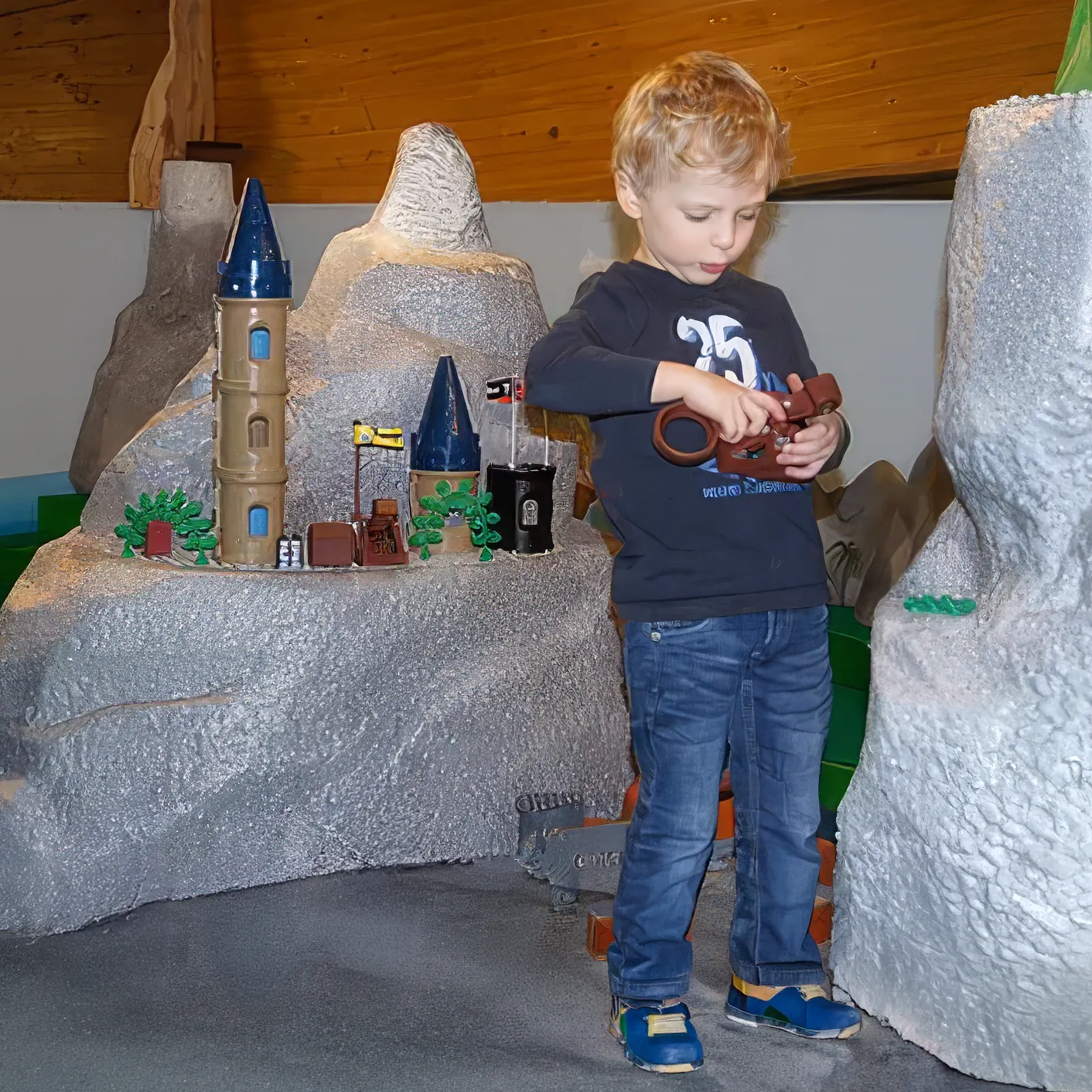 Junge spielt mit braunem Spielzeug vor Modellburg mit Türmen in einer grauen Landschaft.