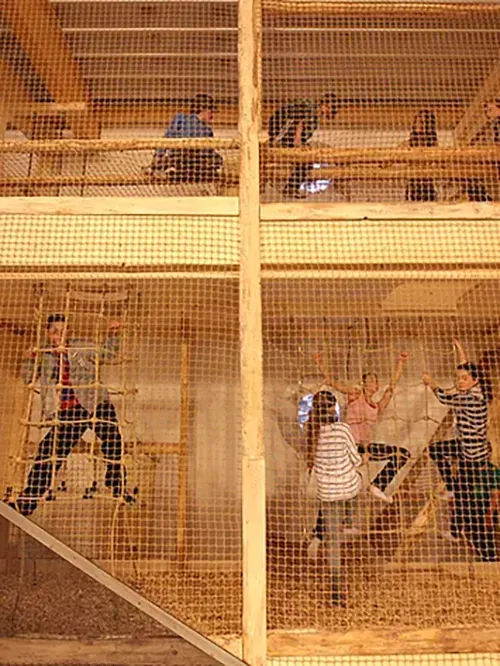 Menschen klettern an einem Netz in einem zweistöckigen Holzgerüst in einer Halle.
